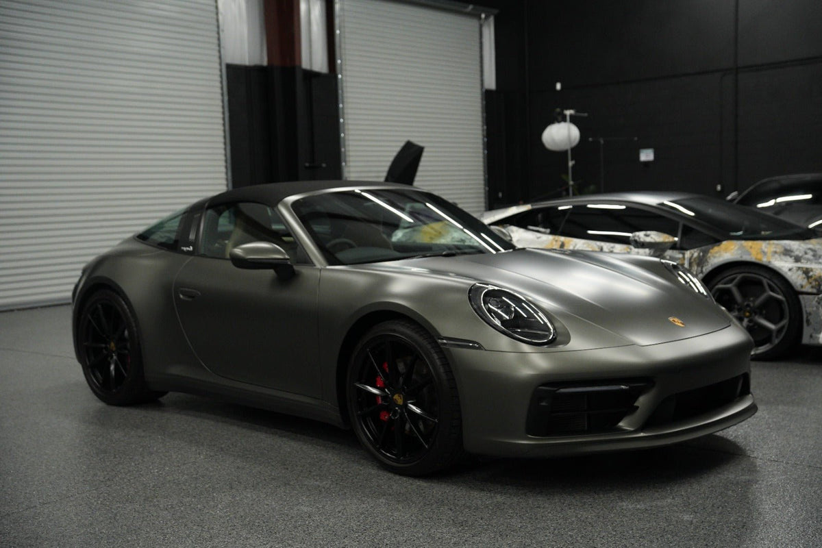 Matte gray Porsche 911 Carrera convertible sports car with black alloy wheels, red brake calipers, and sleek aerodynamic design parked in a garage showroom.