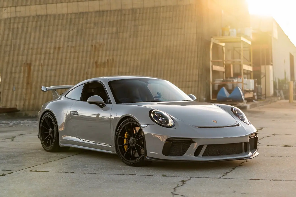 Silver Porsche 911 GT3 sports car with black alloy wheels and rear spoiler, parked on concrete near industrial building in golden hour sunlight.